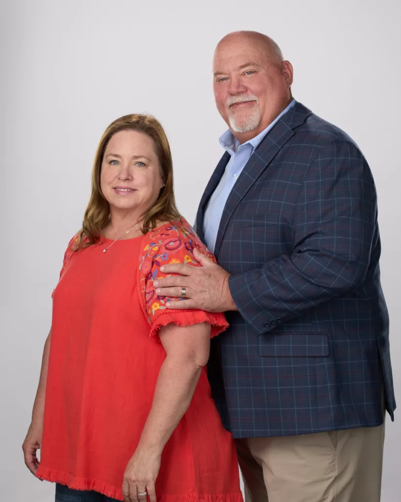 A clear and elegant photo of Jim and his wife, posed side by side against a seamless white backdrop, capturing their shared bond and the simplicity of the setting, highlighting their expressions and attire.