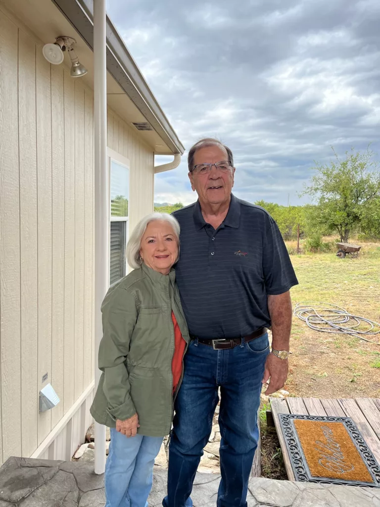 Bill and his wife, standing together and posing in front of a house, reflecting their connection and the context of their residential setting, with details of the home's architecture and landscape in the backdrop.