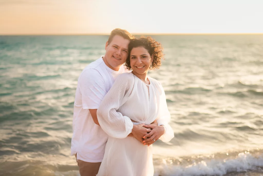 A serene and romantic image of Riley and his wife, posed together at the beach, with the golden hues of the sunset enveloping them, reflecting their bond and the breathtaking beauty of nature.